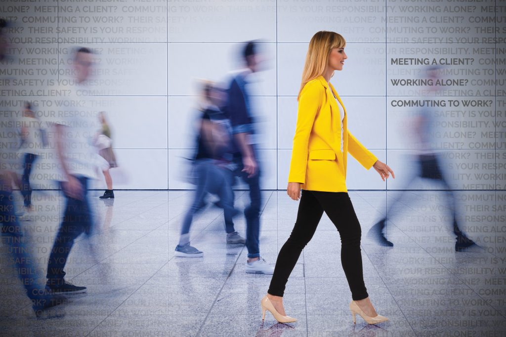 Woman in a bright yellow jacket walking confidently through a busy indoor space, surrounded by blurred figures, highlighting the importance of lone worker safety in dynamic environments for WorkSafe Guardian.